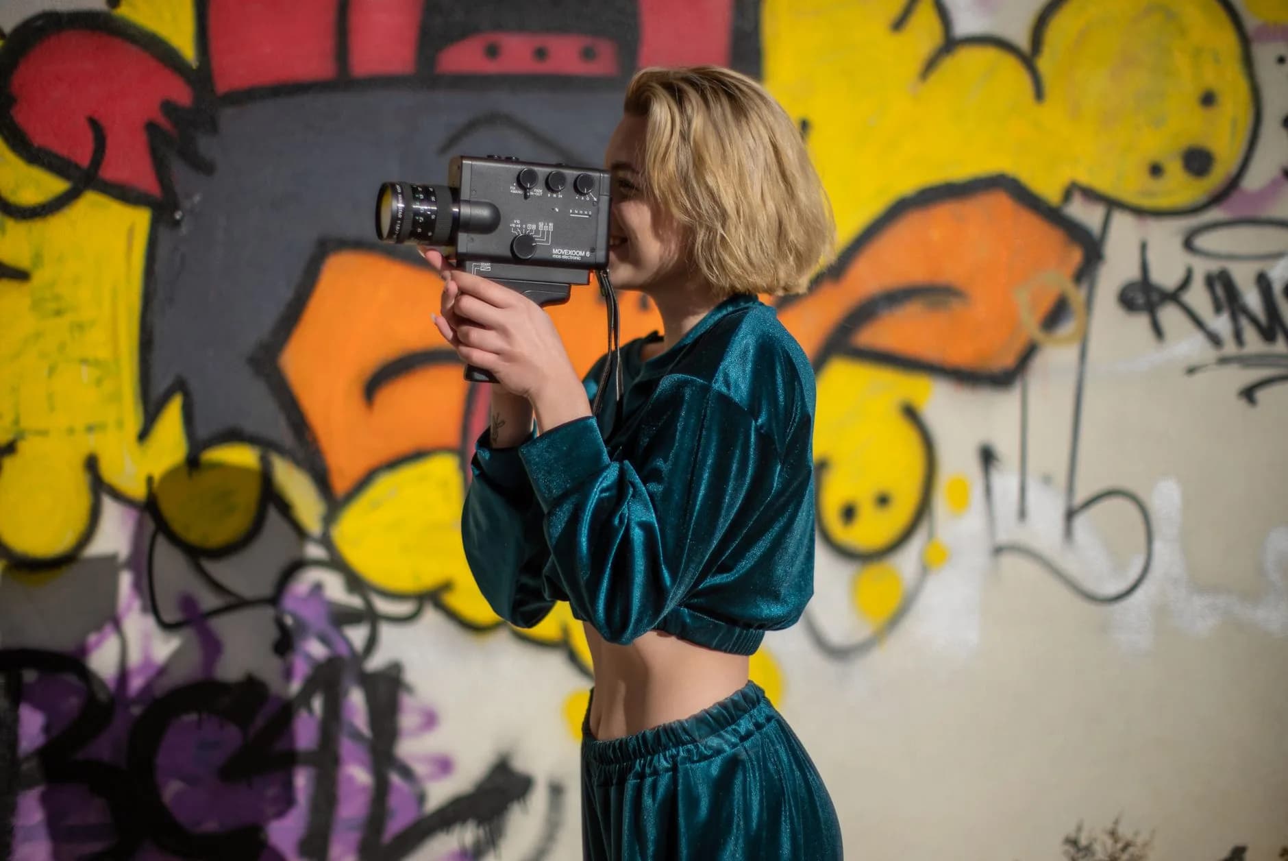 A young woman records video using a vintage camera against a graffiti-covered wall in an urban environment.