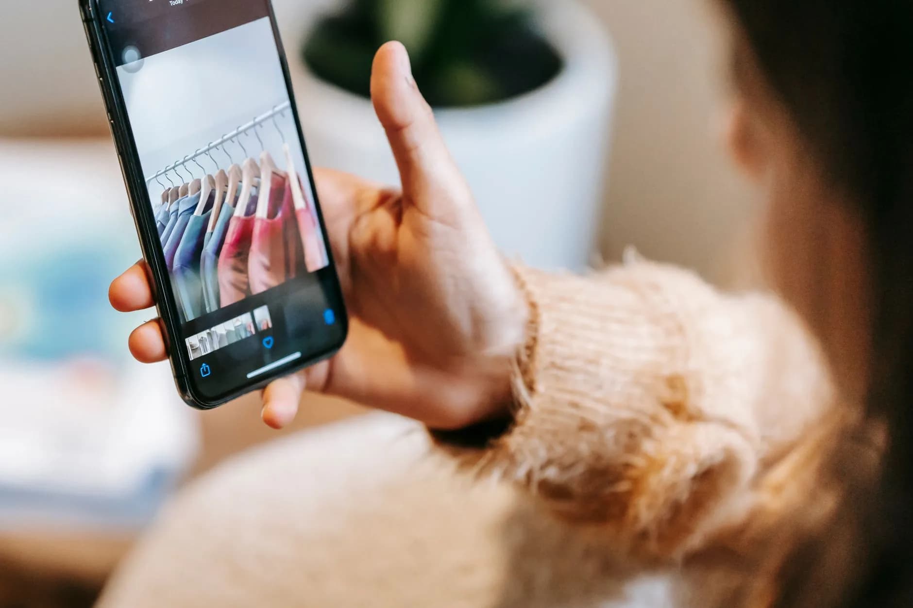 Crop unrecognizable lady in casual clothes looking through pictures of wardrobe on cellphone in light room near potted plant