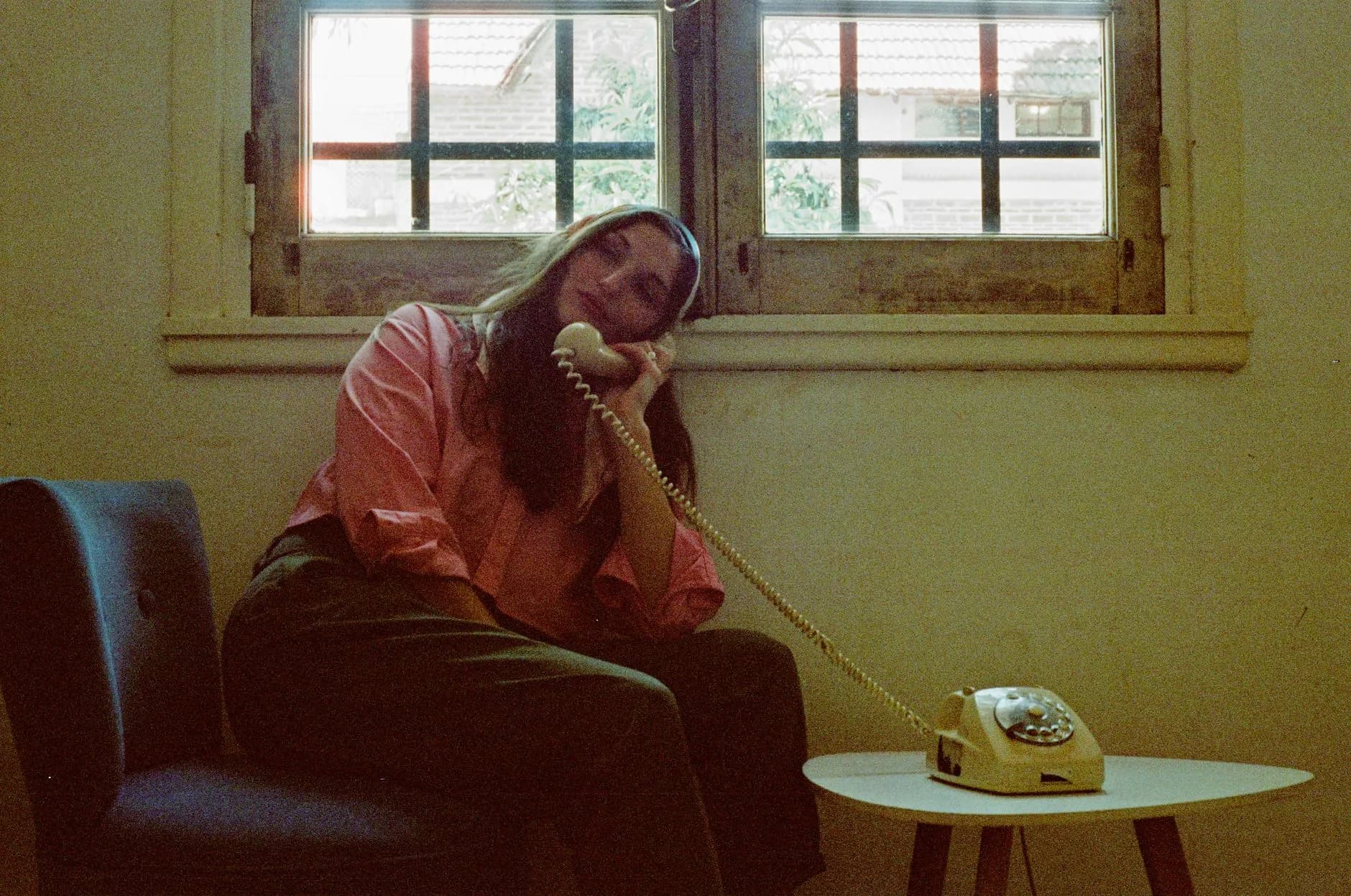 A woman in a pink shirt relaxes on a chair using a vintage rotary phone indoors by a window.